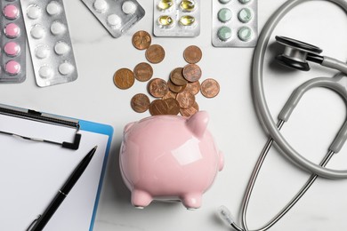 Pale pink ceramic piggy bank with money, stethoscope and pills on white table, flat lay. Medical insurance