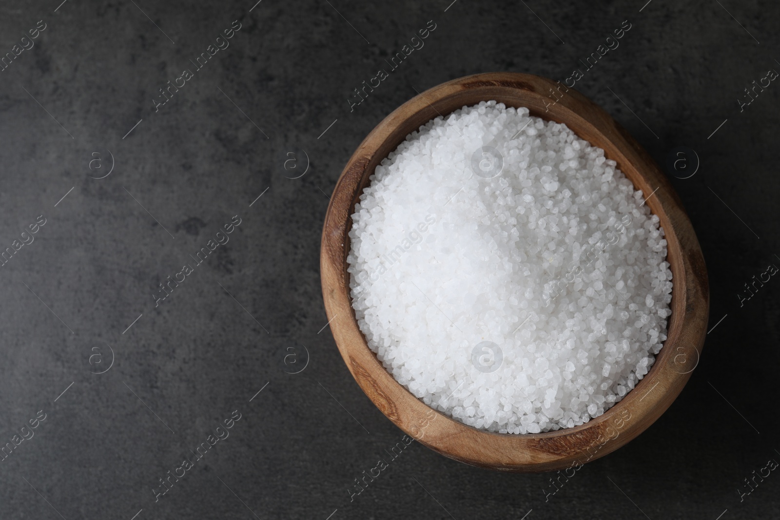 Photo of Natural salt in wooden bowl on dark grey table, top view. Space for text