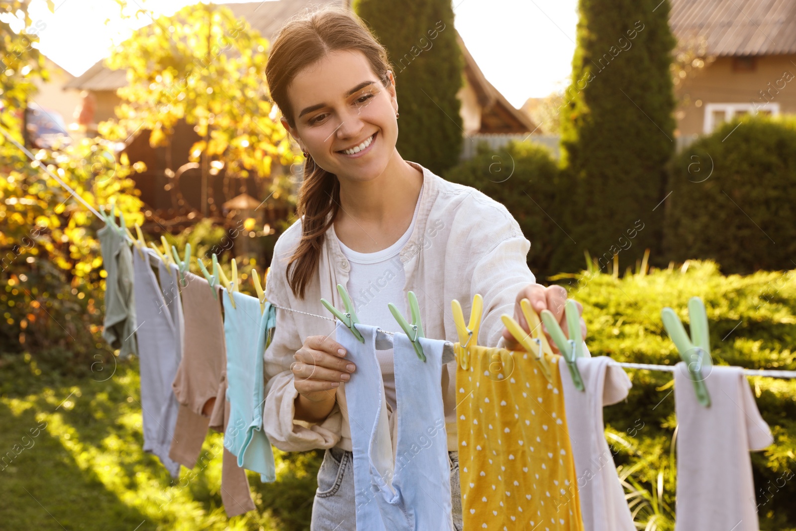 Photo of Smiling woman hanging baby clothes with clothespins on washing line for drying in backyard