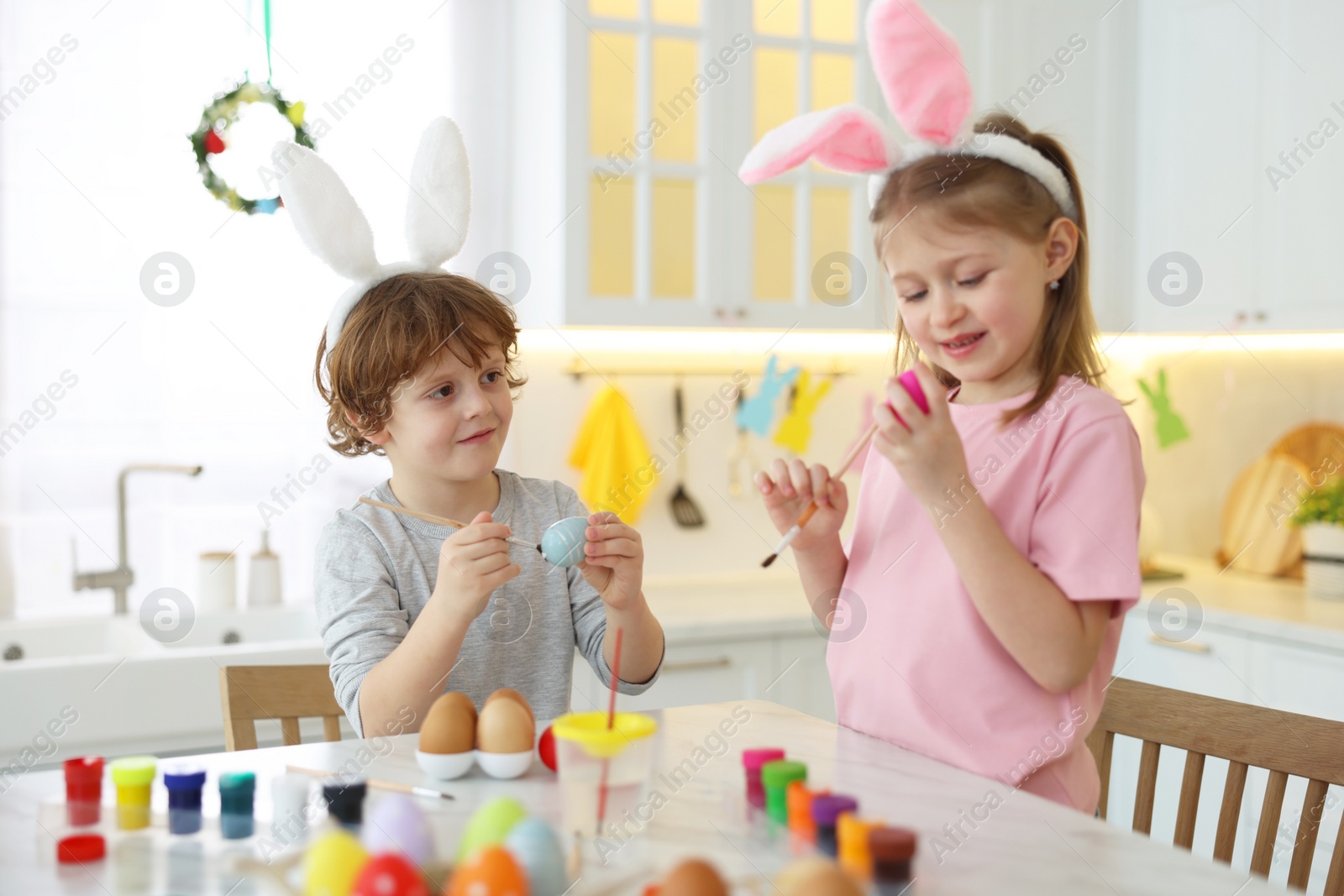 Photo of Easter celebration. Cute children with bunny ears painting eggs at white marble table in kitchen