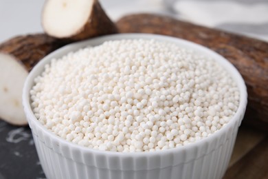 Photo of Tapioca pearls in bowl and cassava roots on table, closeup