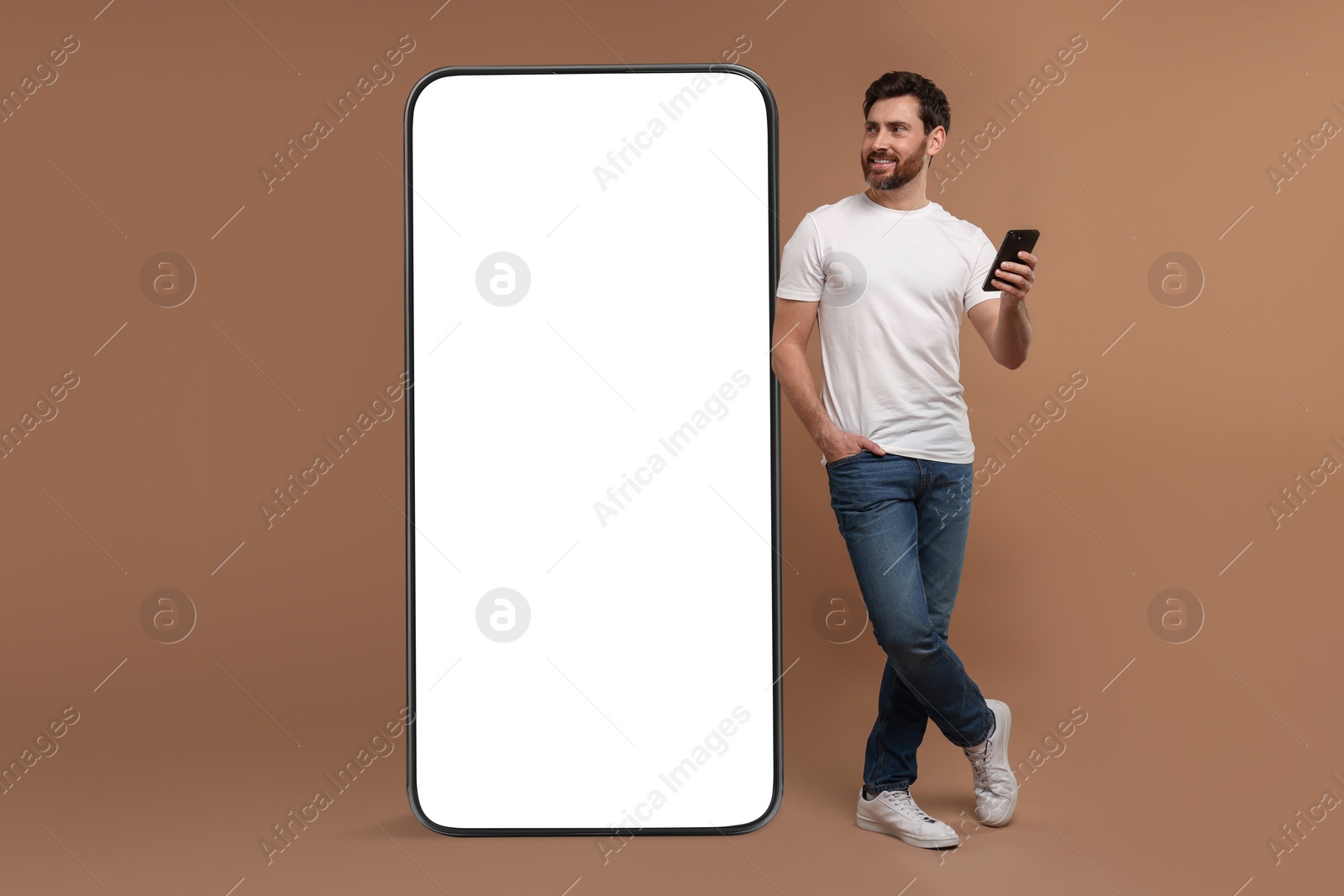 Image of Man with mobile phone standing near huge device with empty screen on dark beige background. Mockup for design