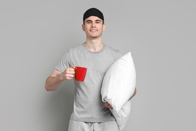 Man in pyjama holding pillow and cup of drink on grey background