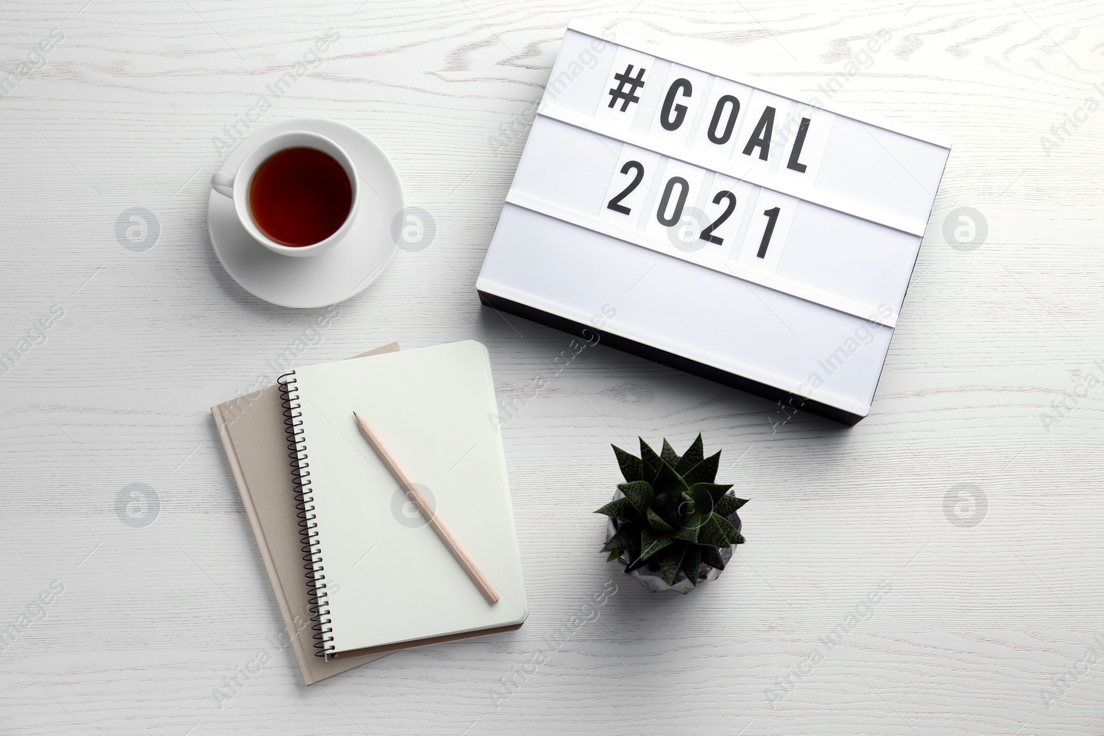 Photo of Light box with hashtag Goal 2021 near notebooks and cup of tea on white wooden table, flat lay. New year targets