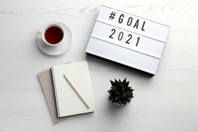 Photo of Light box with hashtag Goal 2021 near notebooks and cup of tea on white wooden table, flat lay. New year targets