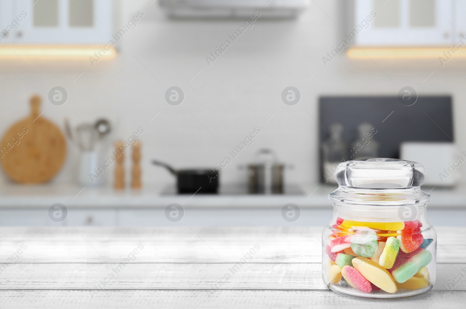 Image of Glass jar with tasty gummy candies on white wooden table in kitchen. Space for text