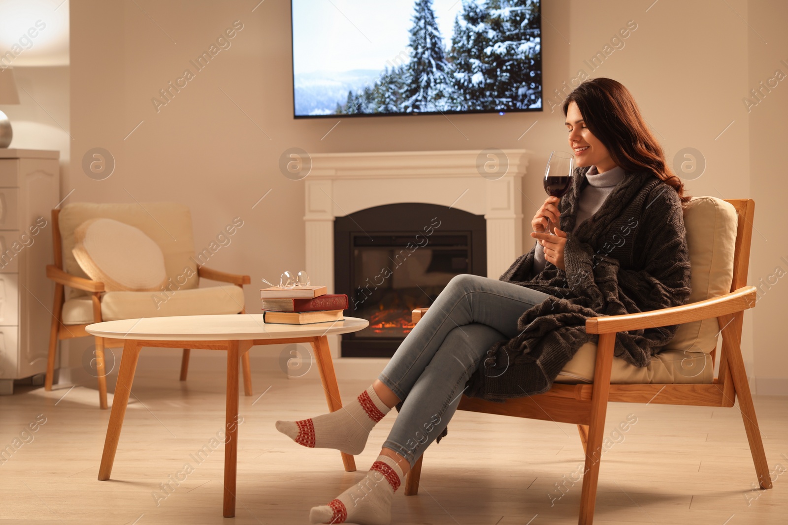 Photo of Young woman with glass of wine resting in armchair near fireplace at home