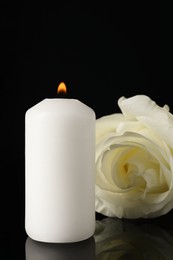 Photo of White rose and burning candle on black mirror surface in darkness, closeup. Funeral symbols