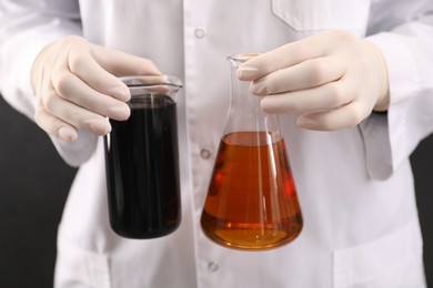 Photo of Woman holding beaker and flask with different types of crude oil on dark background, closeup
