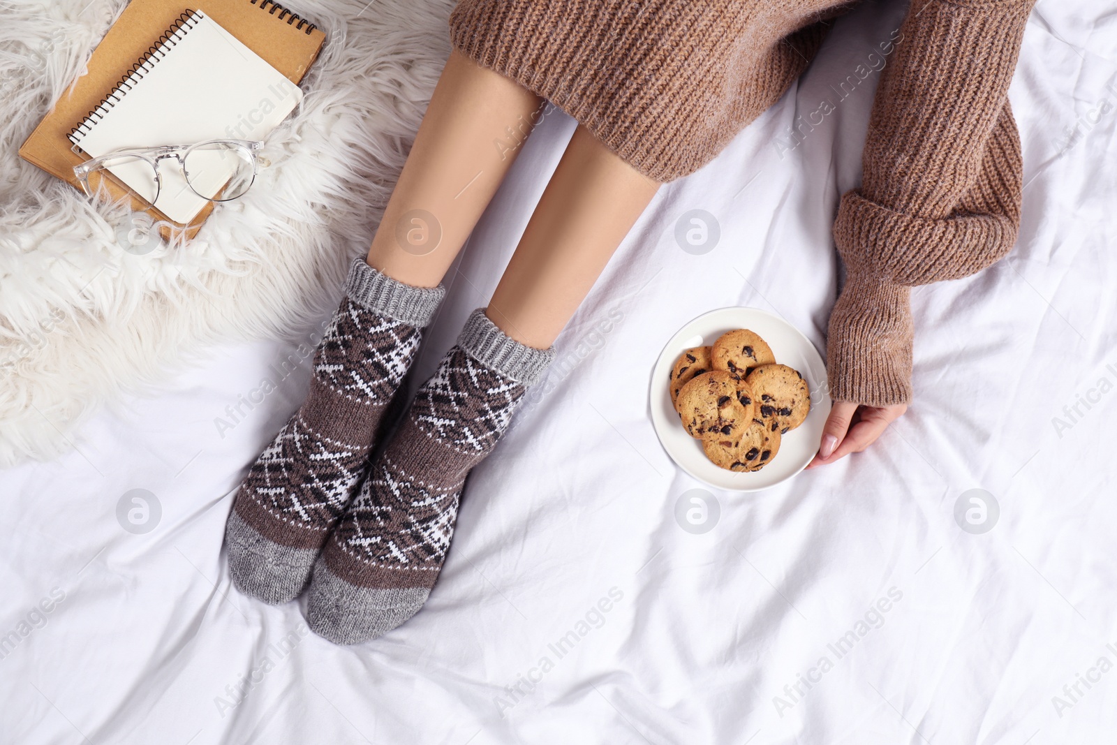 Photo of Woman with cookies wearing knitted socks on white fabric, top view. Warm clothes