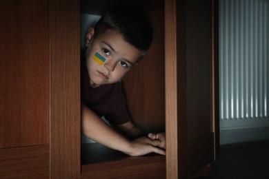 Image of Sad little boy with picture of Ukrainian flag on cheek hiding in wardrobe. Stop war in Ukraine