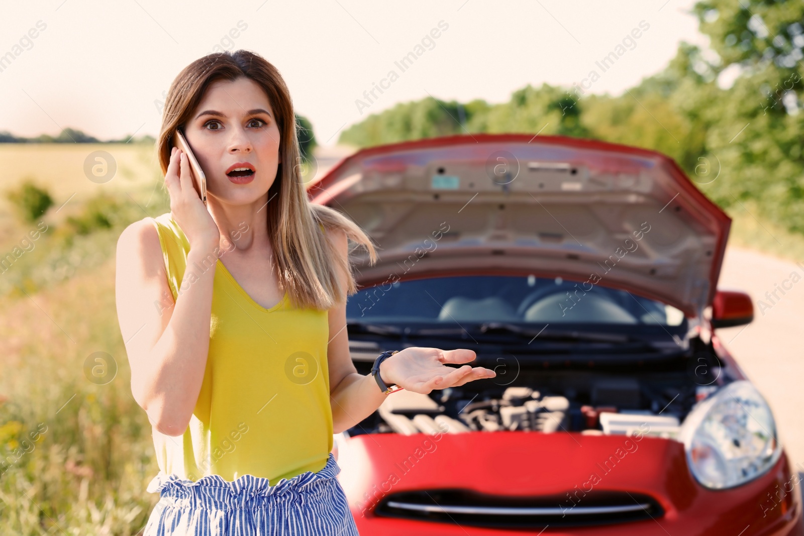 Photo of Stressed woman with mobile phone standing near broken car in countryside