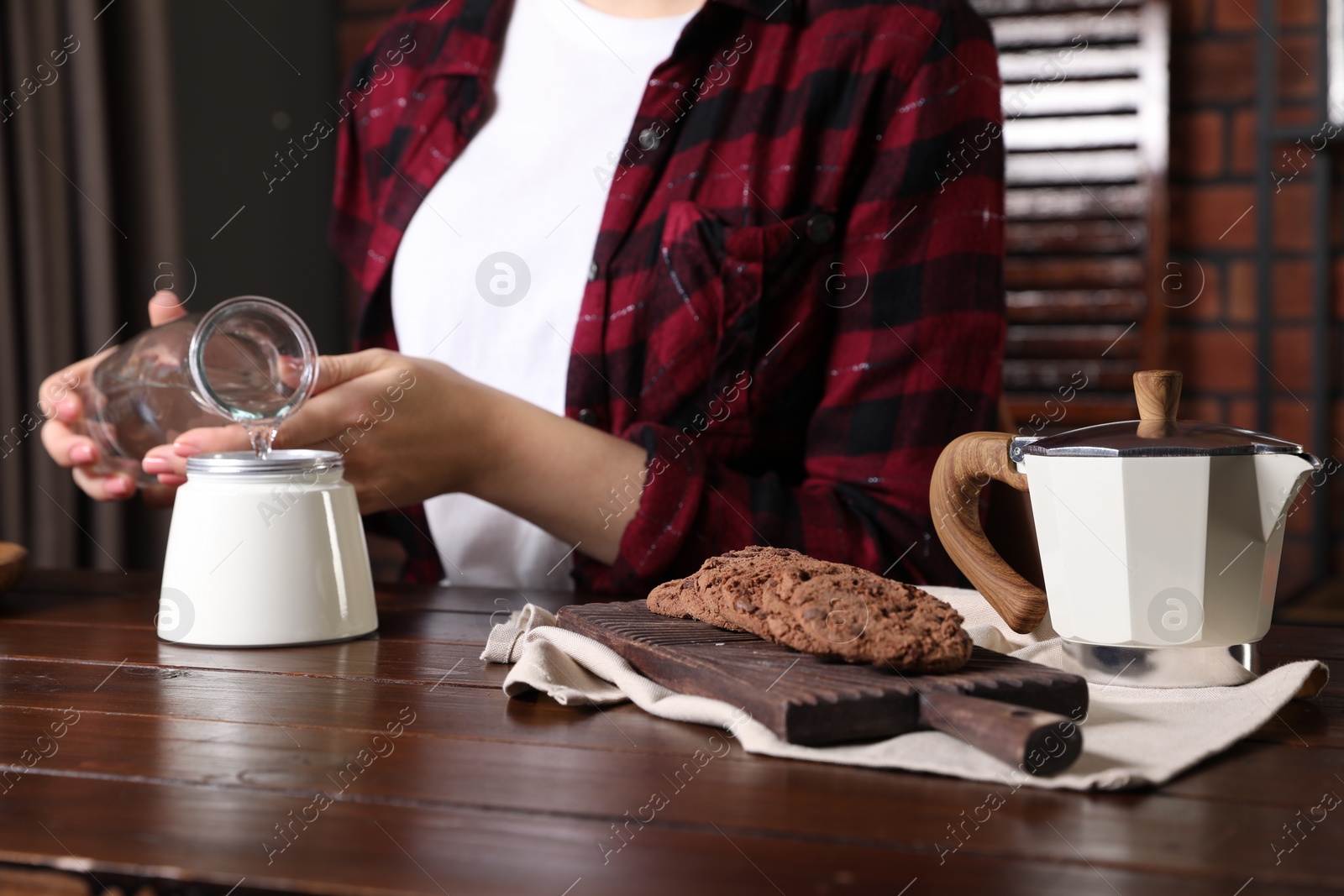 Photo of Brewing coffee. Woman pouring water into moka pot at wooden table indoors, focus on cookies