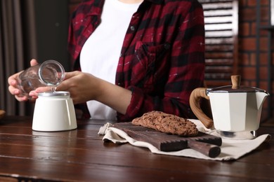 Photo of Brewing coffee. Woman pouring water into moka pot at wooden table indoors, focus on cookies
