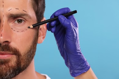 Doctor drawing marks on man's face for cosmetic surgery operation against light blue background, closeup