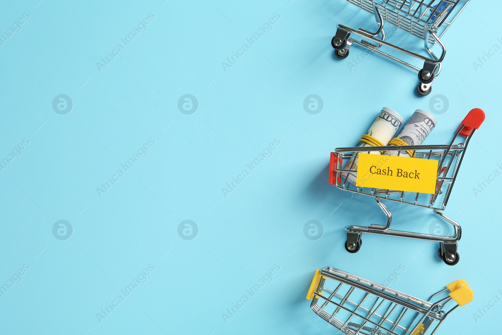 Photo of Card with word Cashback, rolled dollar banknotes and shopping carts on light blue background, flat lay. Space for text