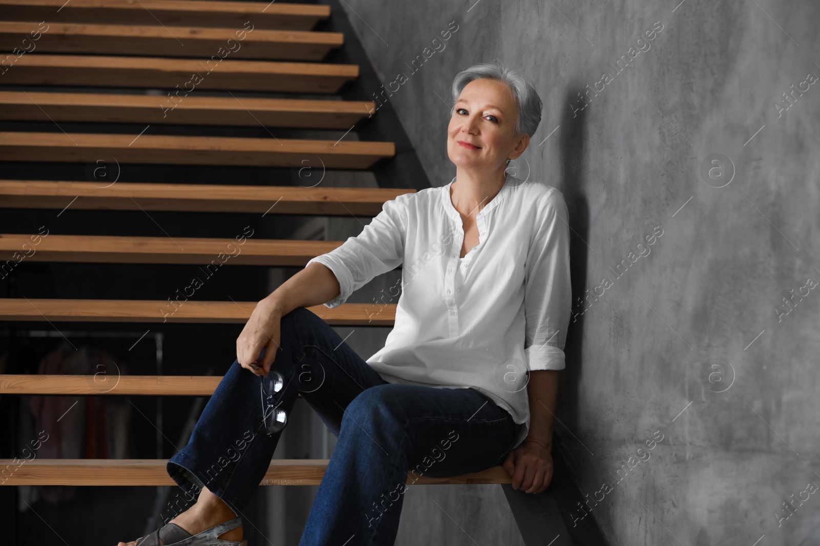 Photo of Happy mature woman sitting on wooden stairs indoors. Smart aging