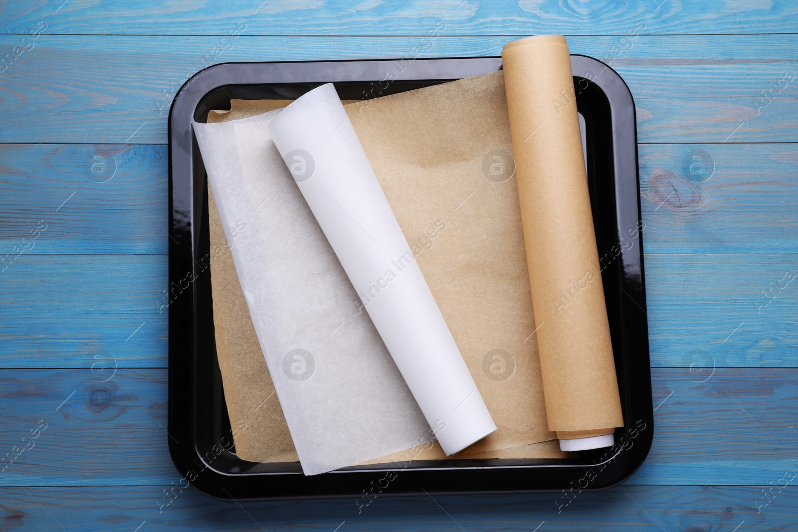 Photo of Rolls of parchment paper in baking pan on light blue wooden table, top view