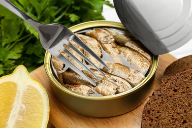 Photo of Fork with canned sprats, lemon and bread on wooden board, closeup
