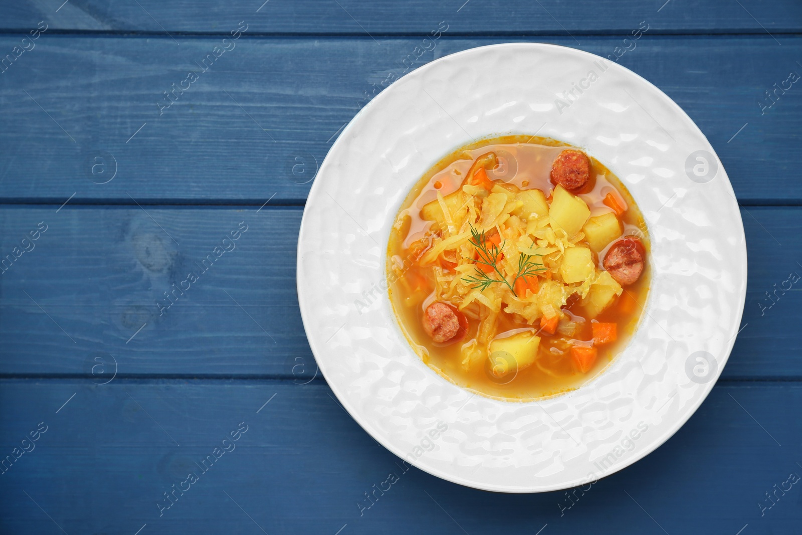 Photo of Delicious sauerkraut soup with smoked sausages and carrot on blue wooden table, top view. Space for text