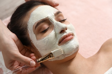 Photo of Cosmetologist applying white mask onto woman's face in spa salon, closeup