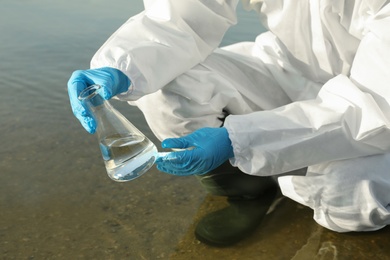 Photo of Scientist in chemical protective suit with conical flask taking sample from river for analysis, closeup