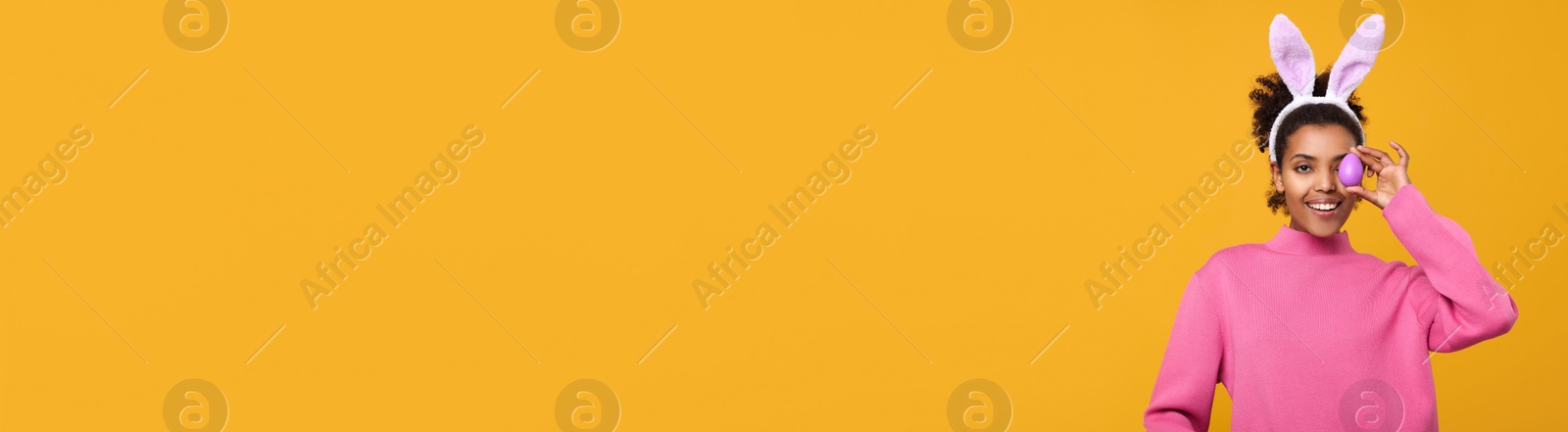 Image of Happy African American woman with bunny ears holding Easter egg near eye on orange background, space for text. Banner design