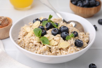 Photo of Tasty oatmeal with blueberries, mint and almond petals in bowl on white tiled table, closeup