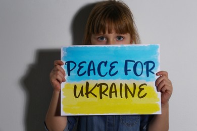 Image of Sad little girl holding paper with phrase Peace for Ukraine near white wall