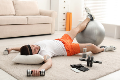 Photo of Lazy young man with sport equipment sleeping on floor at home