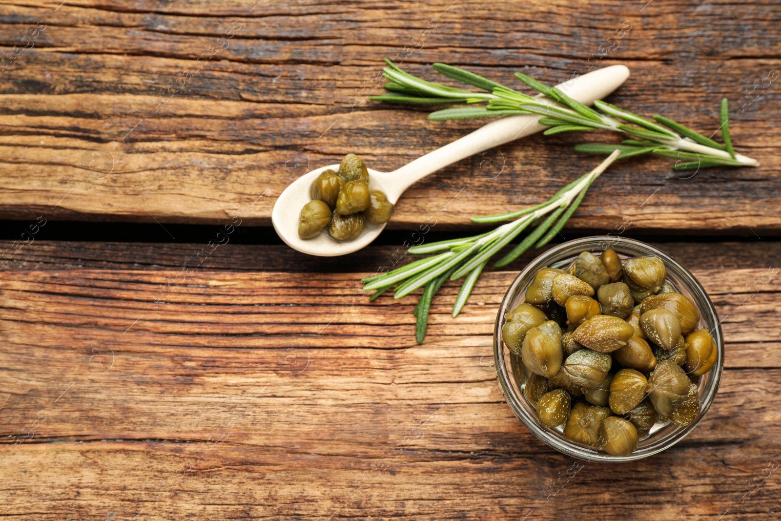 Photo of Tasty capers, rosemary and spoon on wooden table, flat lay. Space for text