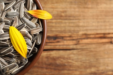 Raw sunflower seeds and petals in bowl on wooden table, closeup. Space for text