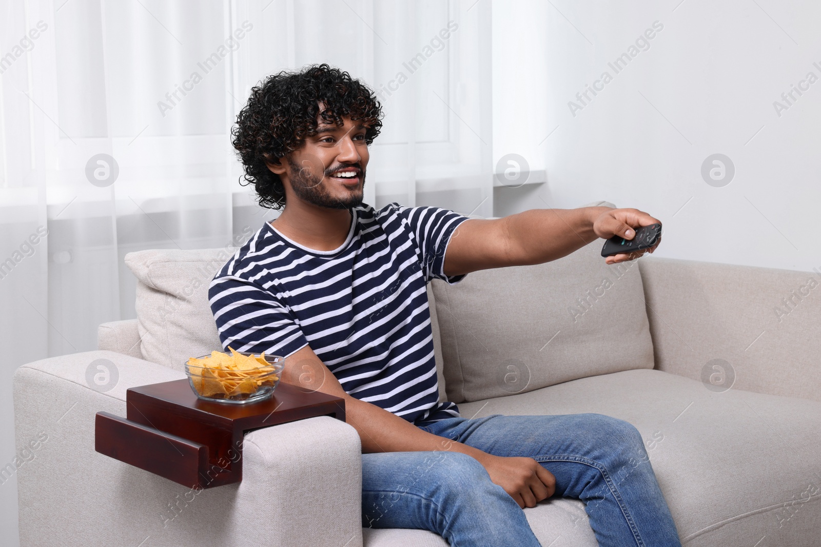 Photo of Happy man switching TV channels at home. Nacho chips on sofa armrest wooden table