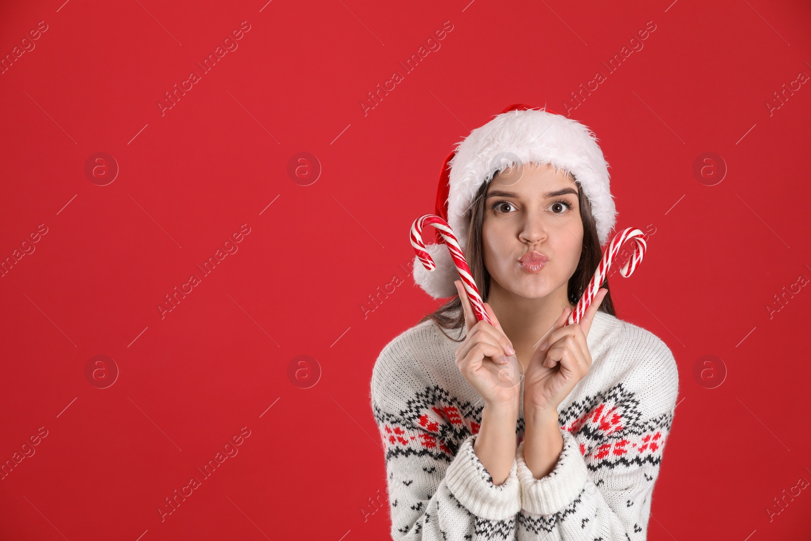 Photo of Pretty woman in Santa hat and Christmas sweater holding candy canes on red background, space for text