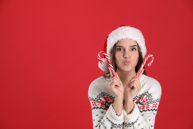 Photo of Pretty woman in Santa hat and Christmas sweater holding candy canes on red background, space for text
