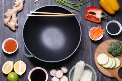 Photo of Empty iron wok and chopsticks surrounded by ingredients on dark grey wooden table, flat lay