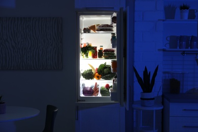 Open refrigerator full of products in stylish kitchen interior at night