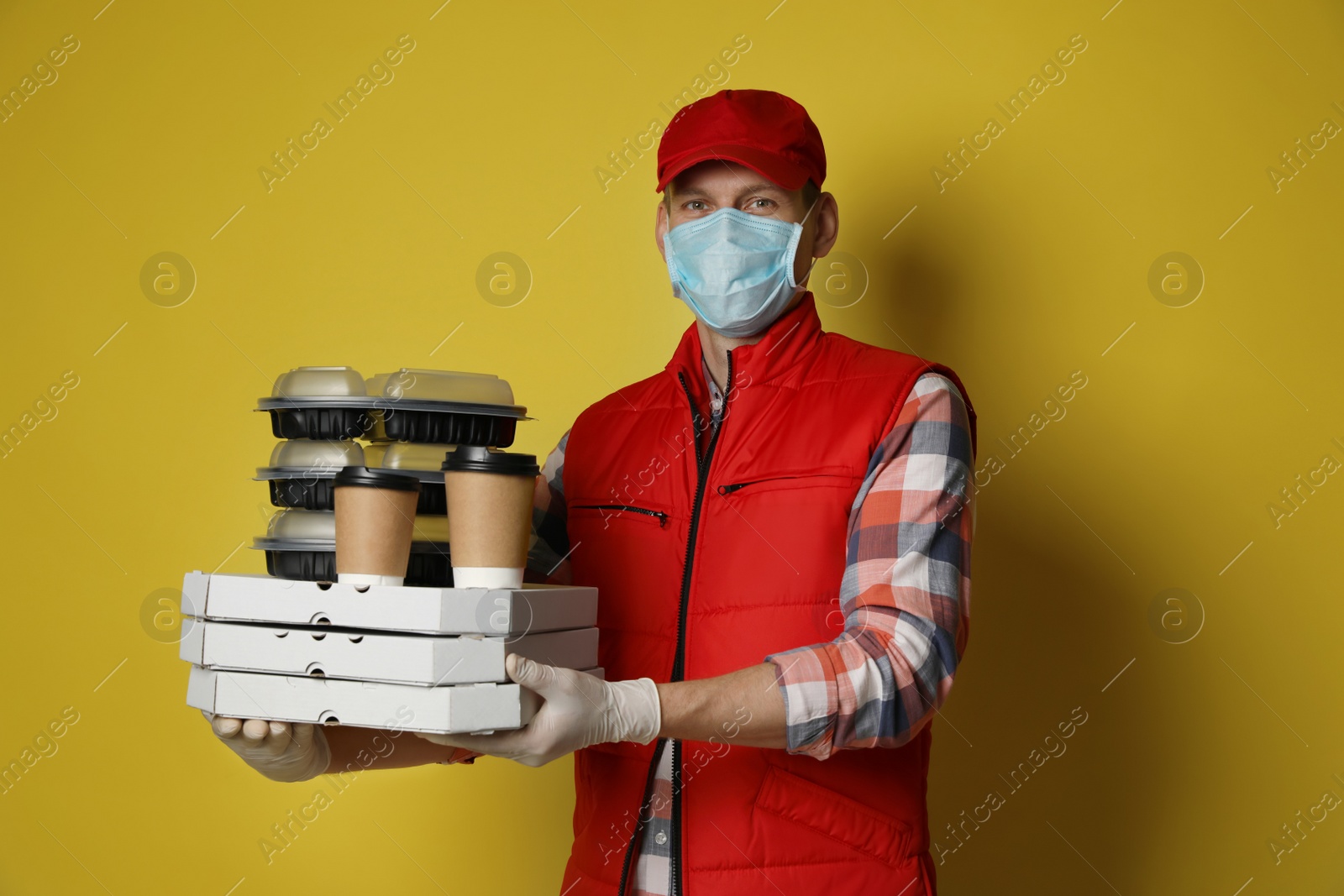 Photo of Courier in protective mask and gloves holding order on yellow background. Food delivery service during coronavirus quarantine