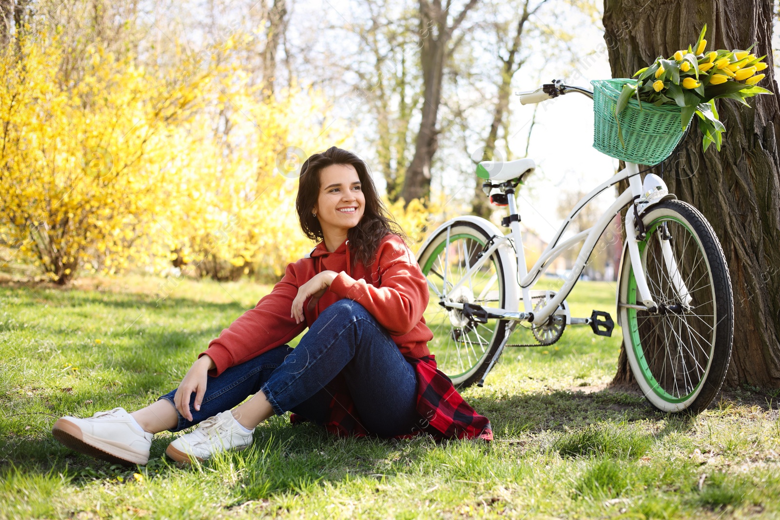 Photo of Beautiful woman sitting on green grass near bicycle with bouquet of yellow tulips outdoors. Spring day