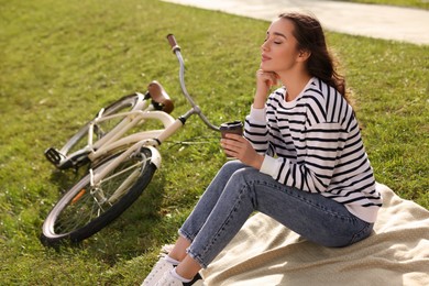 Photo of Young woman sitting on green grass and holding cup of coffee near bicycle outdoors, space for text