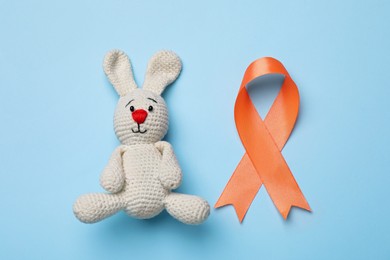 Photo of Orange ribbon and toy bunny on light blue background, flat lay. Multiple sclerosis awareness