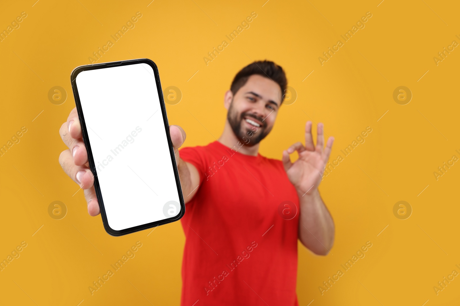 Photo of Young man showing smartphone in hand and OK gesture on yellow background, selective focus. Mockup for design