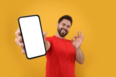 Photo of Young man showing smartphone in hand and OK gesture on yellow background, selective focus. Mockup for design