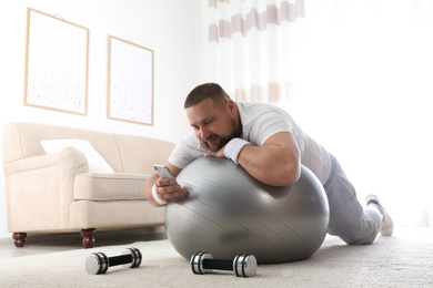 Photo of Lazy overweight man using smartphone while lying on exercise ball at home
