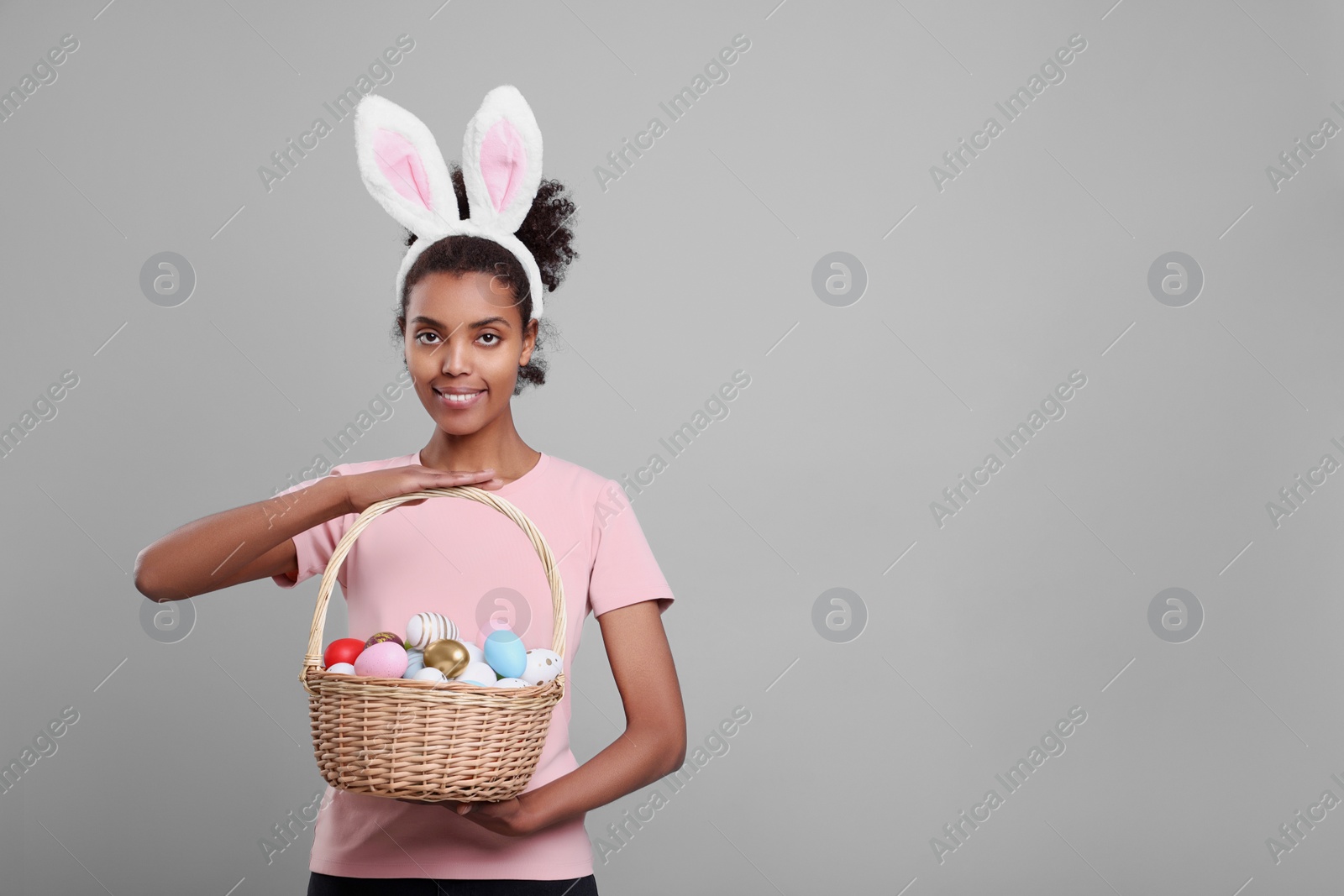 Photo of Happy African American woman in bunny ears headband holding wicker basket with Easter eggs on gray background, space for text