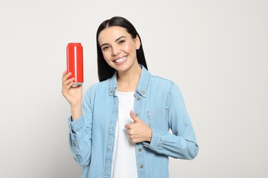 Beautiful happy woman holding red beverage can and showing thumbs up on light grey background