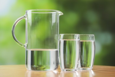 Photo of Jug and glasses with clear water on table against blurred green background, closeup