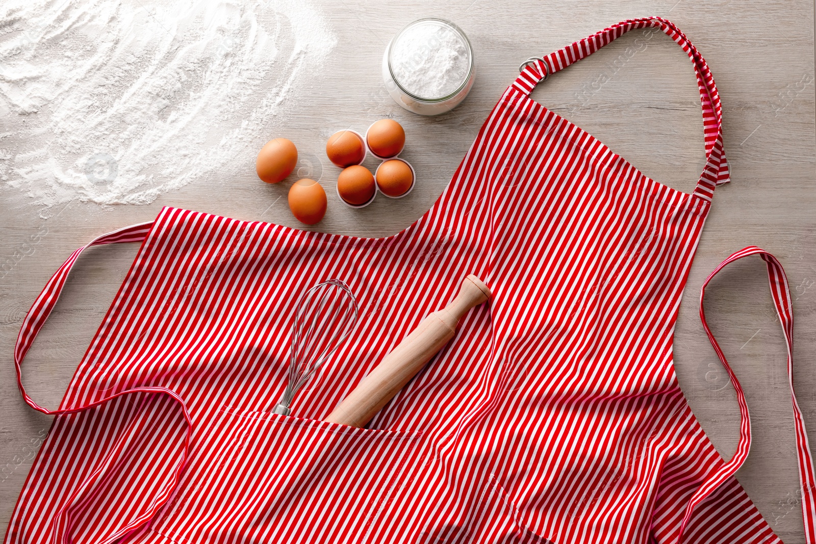 Photo of Red striped apron with kitchen tools and different ingredients on wooden table, flat lay
