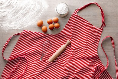 Red striped apron with kitchen tools and different ingredients on wooden table, flat lay
