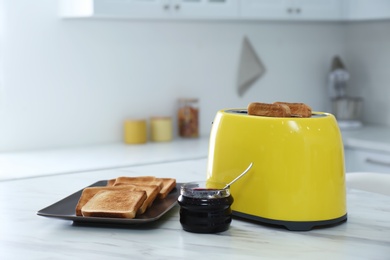 Photo of Modern toaster with slices of bread and jam on white marble table in kitchen